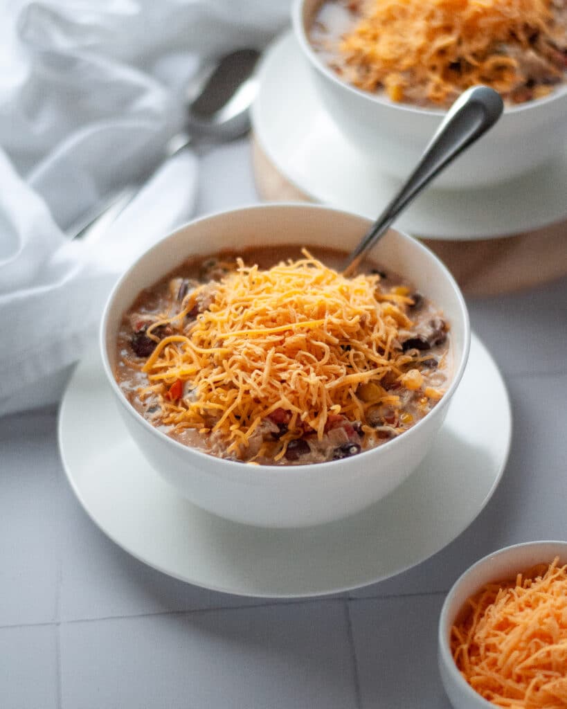 A large bowl filled with crockpot white chicken chili topped with cheese. The bowl has a spoon in it. Another bowl of chili sits in the background and a small bowl of cheese in the front.