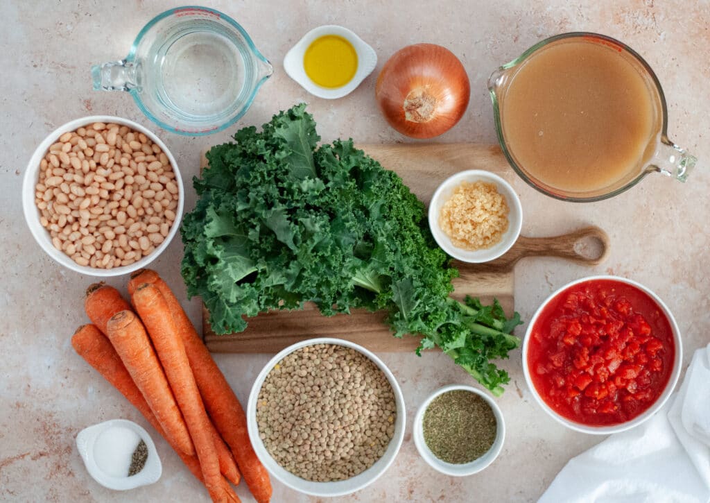Ingredients for Tuscan white bean soup. This top down photo includes white beans, lentils, kale, diced tomatoes, carrots, chicken bone broth, water, olive oil, onion, garlic, Italian seasoning, salt, and pepper, artfully arranged.