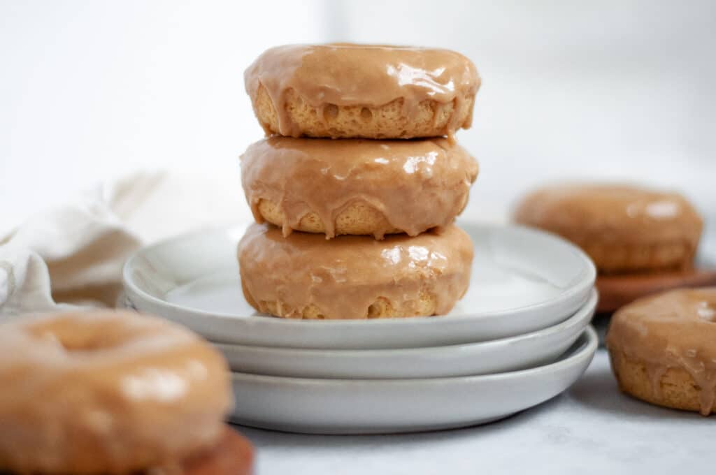 Stack of maple baked donuts on a stack of plates. The maple donuts stack is surrounded by additional baked maple donuts in the foreground and background.