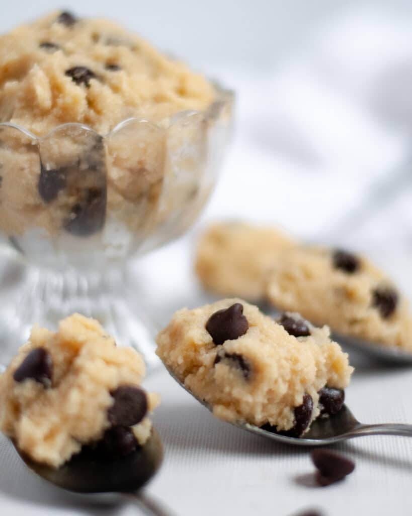 four spoons around a cute glass serving dish, all filled with the gluten free edible cookie dough with chocolate chips. A few chocolate chips are scattered around the spoons and a white linen is in the background.