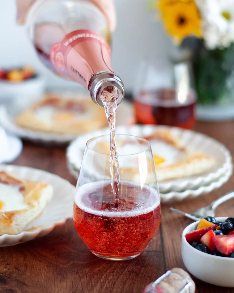 A stop motion image of bottle of Taittinger rose champagne being poured into a stemless wine glass is the focal point of this image. Around the drink are the puff pastry egg tars, fruit salad, forks, and a vase of yellow and white flours.