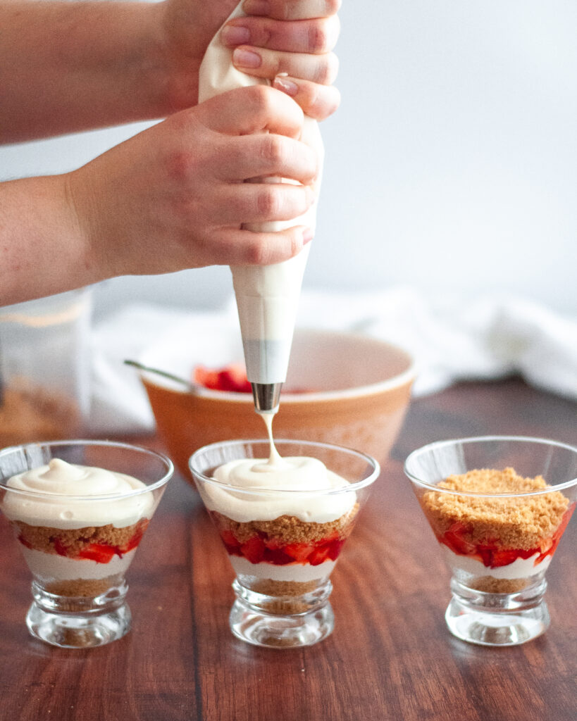 A line of three stemless martini glasses being filled with the various layers of this deconstructed cheesecake recipe. Two hands are holding a piping bag filled with cheesecake filling and the person has just finished filling the middle glass with the second cheesecake layer.