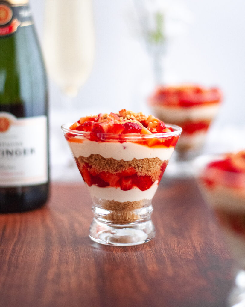 close up of a glass filled with the strawberry cheesecake cups recipe. additional cups are in the foreground and background. there is also a bottle of taittinger champagne and a glass of champagne in frame too.