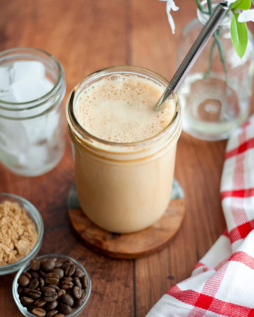 A completed Iced Brown Sugar Oatmilk Shaken Espresso in a mason jar with a metal reusable straw. The jar is sitting on a coaster and surrounded by a jar of ice, a bowl of brown sugar, a bowl of coffee beans, a red and white linen, and a vase of white flowers.