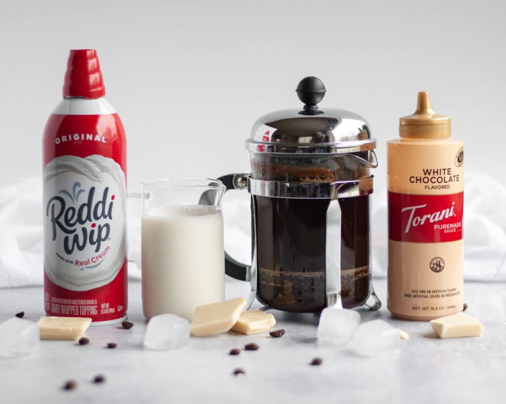 Ingredients needed to make a white chocolate iced cafe mocha. This includes a can of whipped cream, milk, strong brewed coffee, and white chocolate sauce.