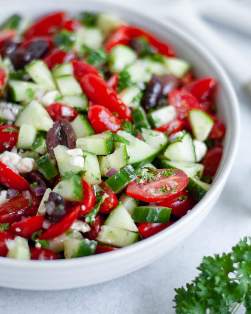 close up view of a bowl filled with tomato cucumber feta salad with Kalamata olives, parsley, onions, and an light oil dressing.