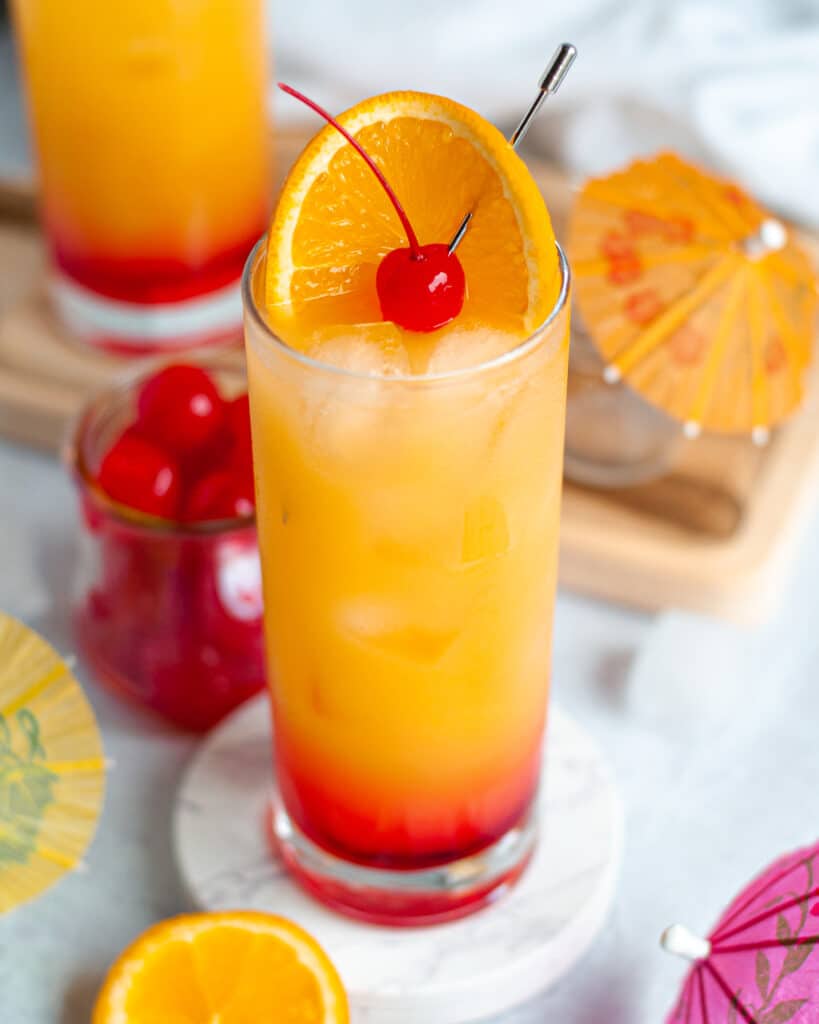 Close up of a highball filled with this vodka sunrise recipe and garnished with an orange slice and maraschino cherry. The glass is surrounded by drink umbrellas, more cherries, with another vodka sunrise cocktail in the background.