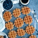 Top-down view of whole wheat waffles cooling on a wire rack with a bowl of blueberries and two forks nearby.
