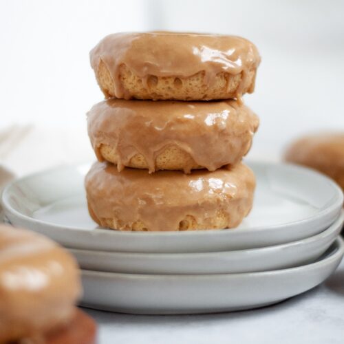 Tabletop shot of three baked maple donuts with maple glaze on a stack of three white plates.