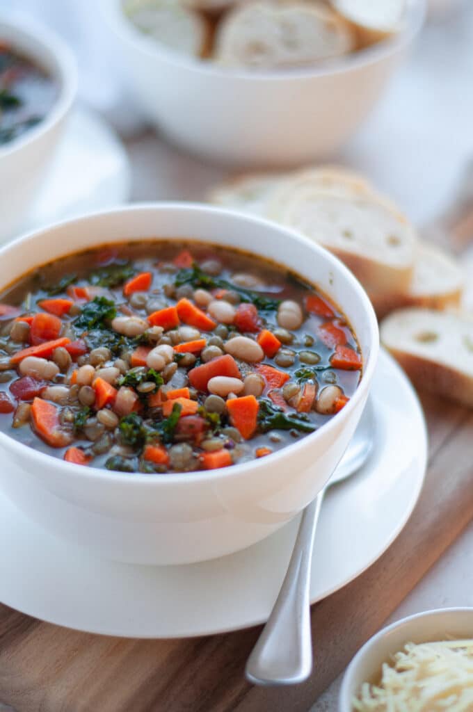 Close up of a bowl filled with this Tuscan white bean soup with kale and lentils. This high-protein collagen soup is surrounded by slices of bread, a bowl of shredded parmesan, and another bowl of chicken bone broth soup.
