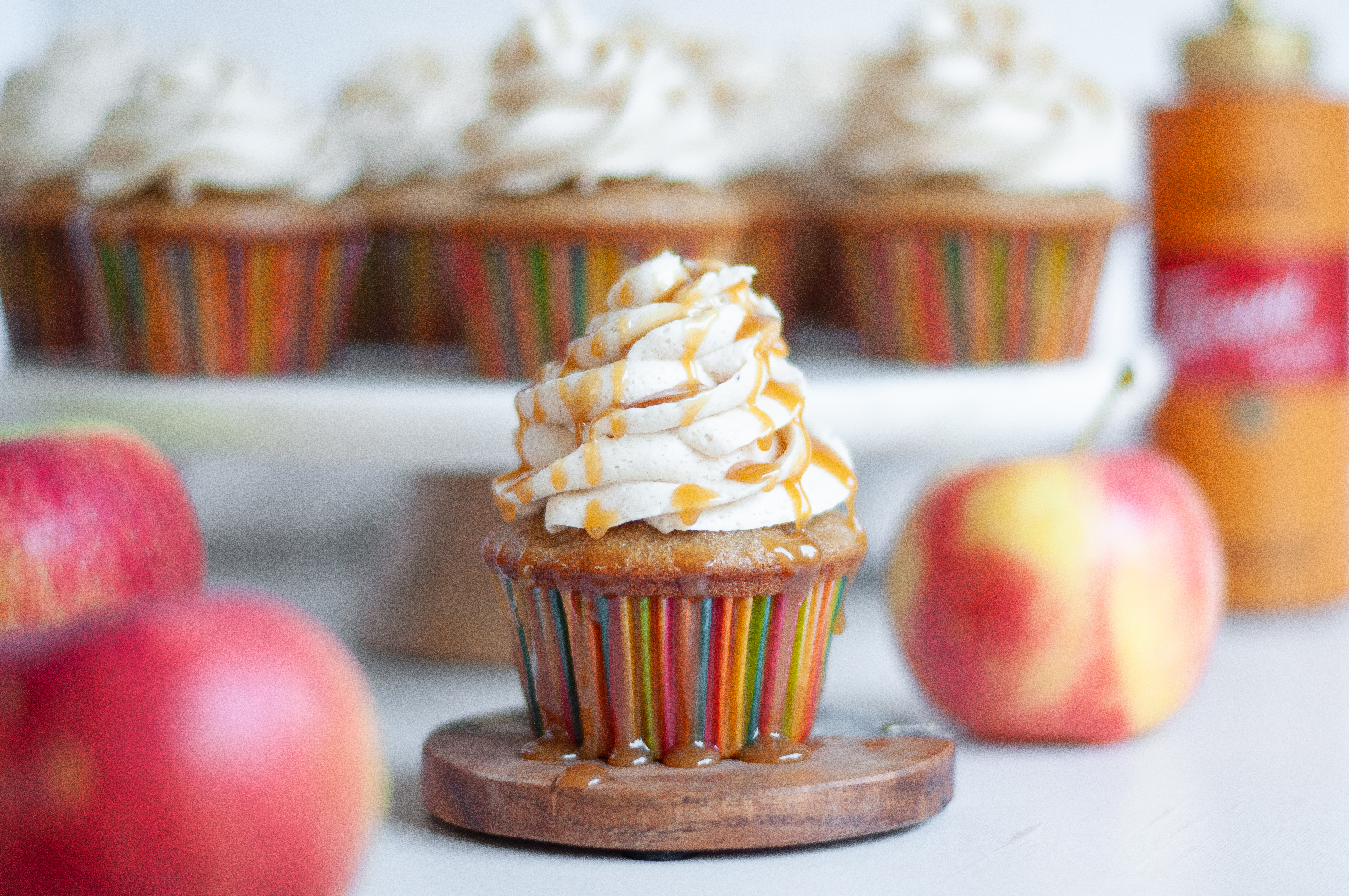 Close up of an apple spice cupcake with spiced buttercream frosting and caramel drizzle. Behind the cupcake in focus is a cake stand covered with more spiced cupcakes and a jar of caramel syrup. The apple cupcake is also surrounded by apples.