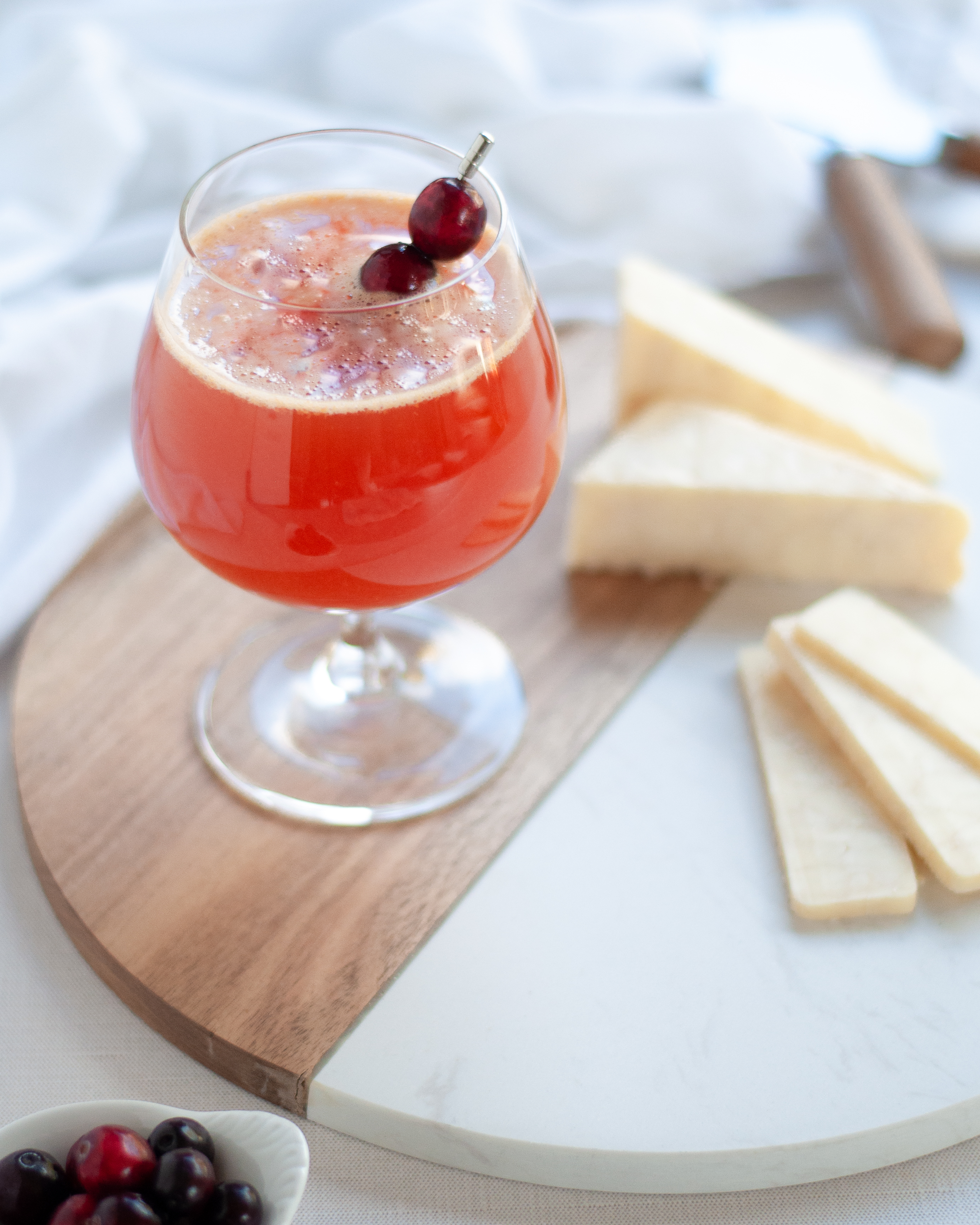 A cranberry shandy garnished with fresh cranberries is sitting on a serving board with sliced gourmet cheese.