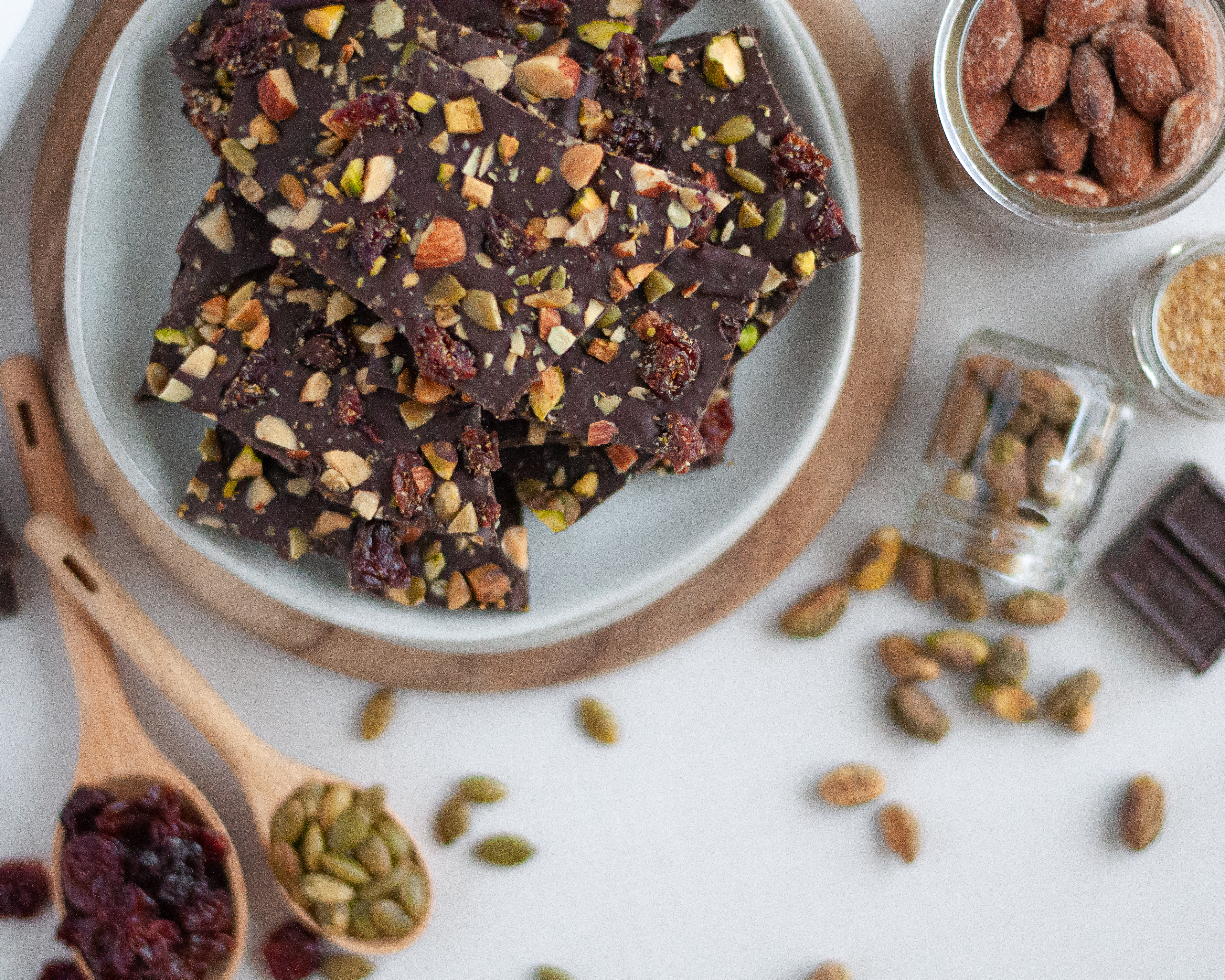 Top down view of a plate of dark chocolate bark with fruit and nuts, surrounded by the ingredients for chocolate bark: chocolate, ground flax, almonds, pistachios, pepitas, and cherries.