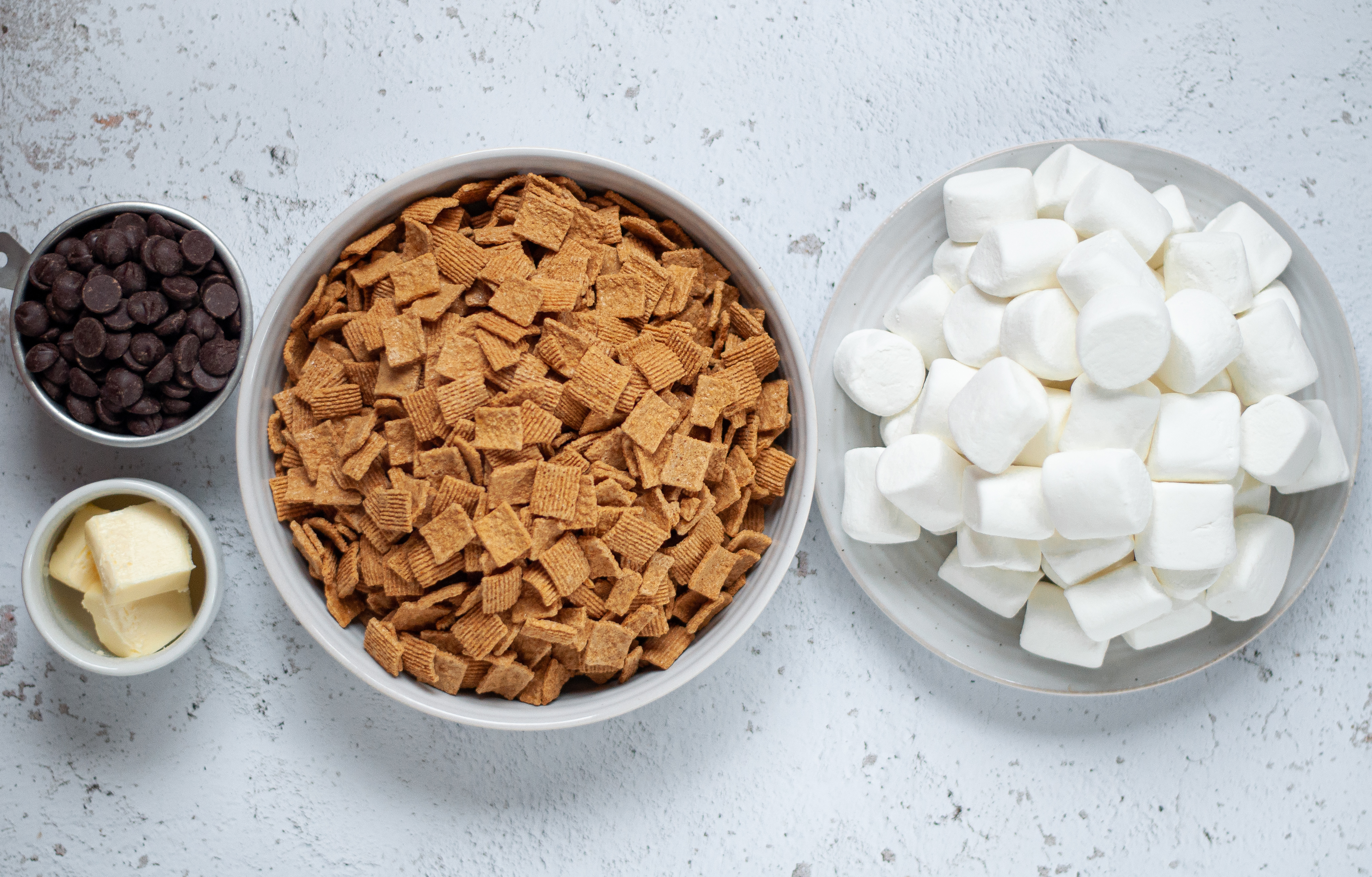 Ingredient shot for this cereal bars recipe. Includes a measuring cup filled with chocolate chips, 3 tablespoons of butter in a small bowl, a large bowl of graham cereal, and a large plate with jet-puffed marshmallows.