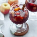 close up shot of two wine glasses filled with fall red wine sangria and garnished with a cinnamon stick. the pitcher of sangria is in the background, with apples and a white linen.