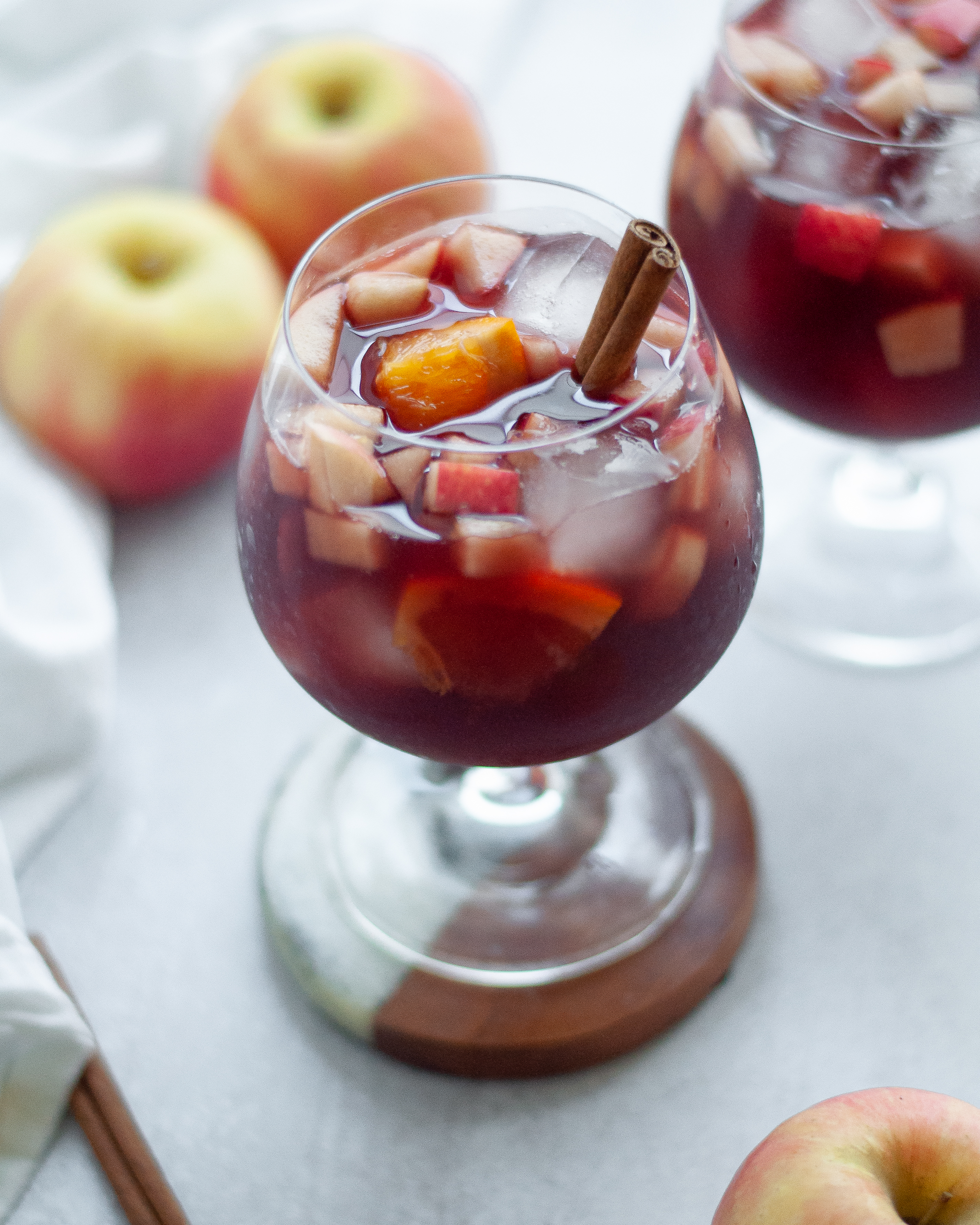Close up view of this fall sangria recipe, garnished with a cinnamon stick. There is another glass of fall red wine sangria in the background, apples, cinnamon sticks, and a white napkin in the photo as well.