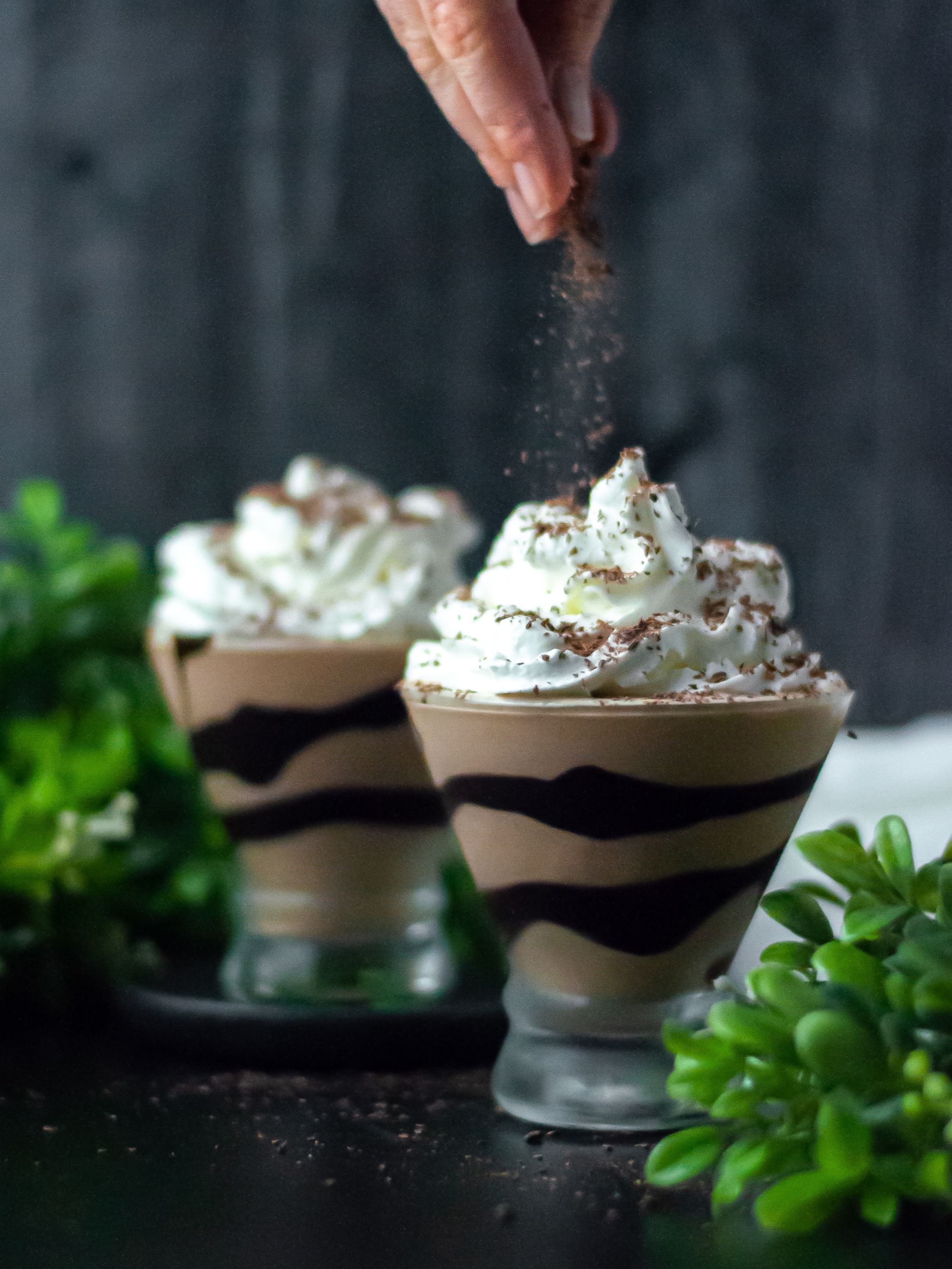 Two glasses of frozen mudslides, topped with whipped cream, and in the process of being sprinkled with chocolate shavings.