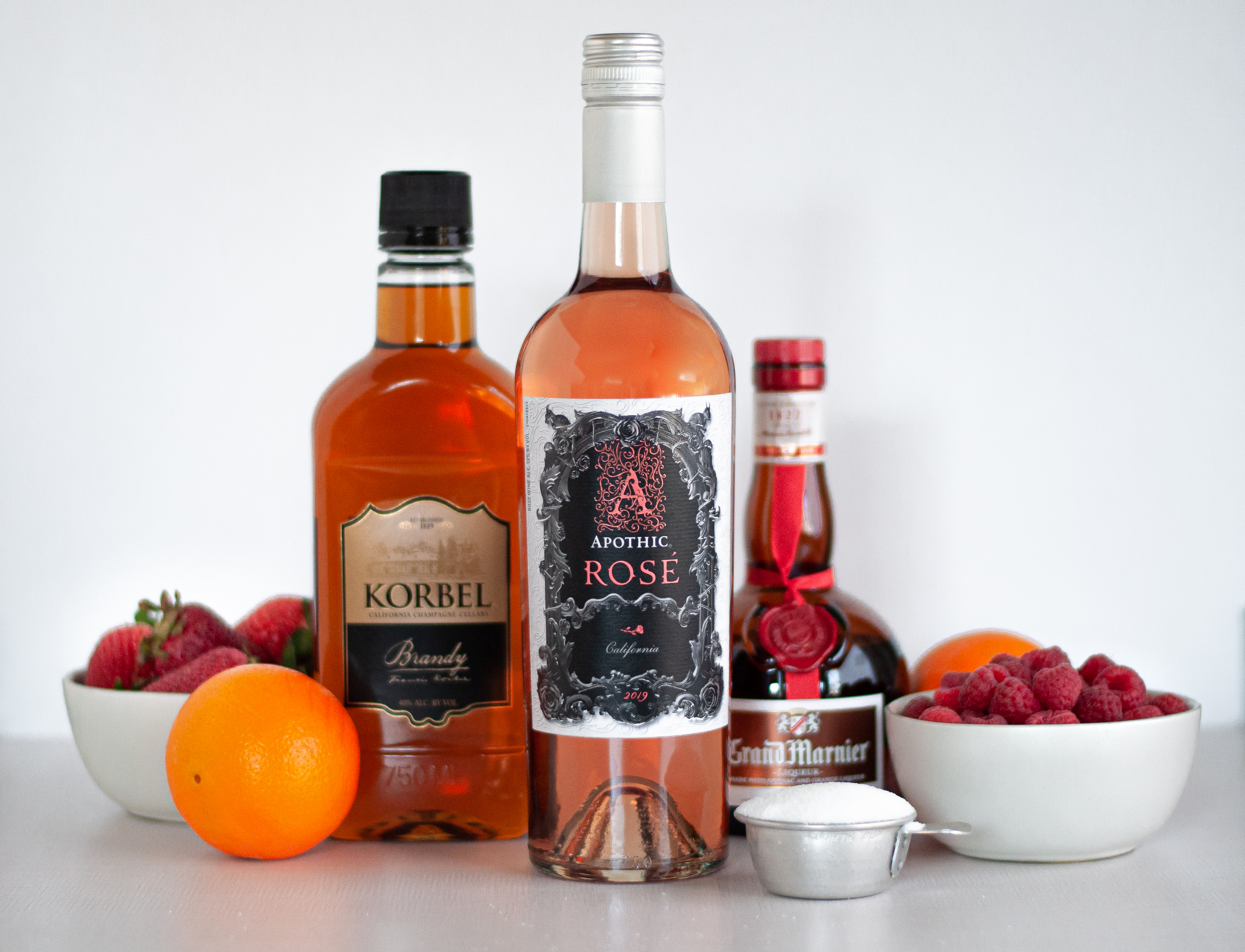Ingredients for rosé sangria, including a bottles of rosé wine, Korbel brandy, and Grand Marnier. A 1/4 cup measure filled with granulated sugar, 2 oranges, and bowls of strawberries and raspberries.