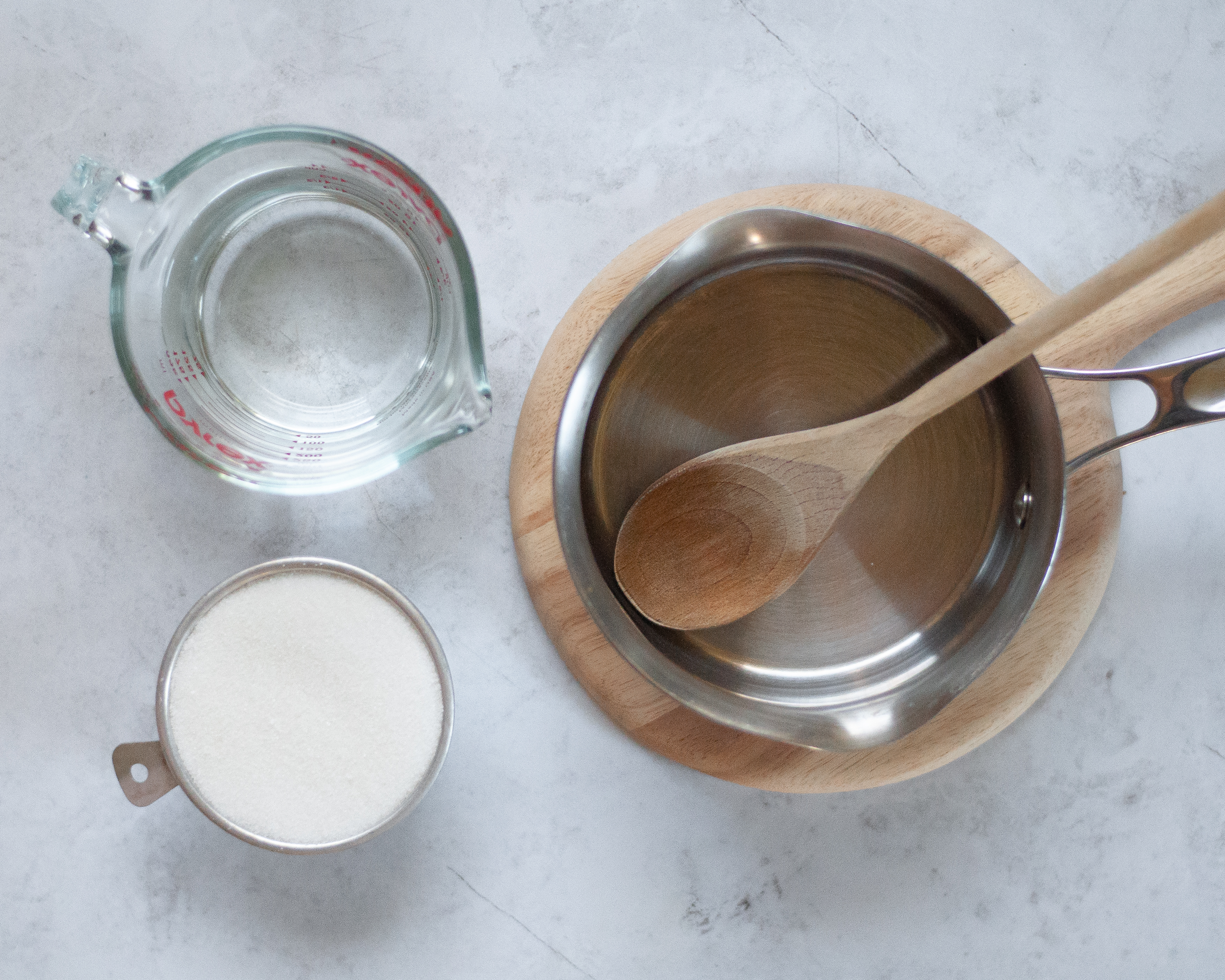 Ingredients and equipment to make homemade simple syrup. Includes water, sugar, a small saucepan, and a wooden spoon.