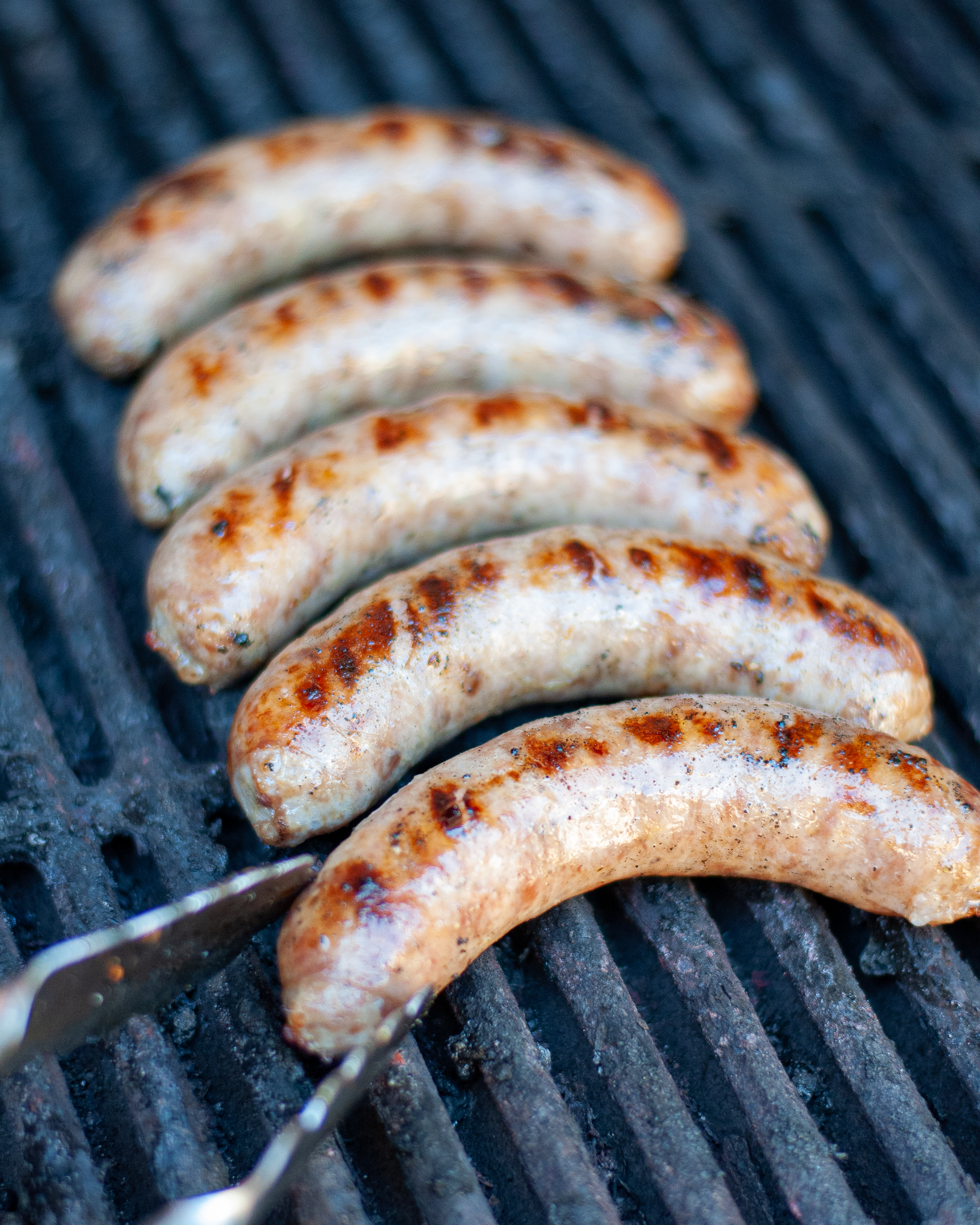 cooking brats on grill, with a tongs ready to turn a brat.