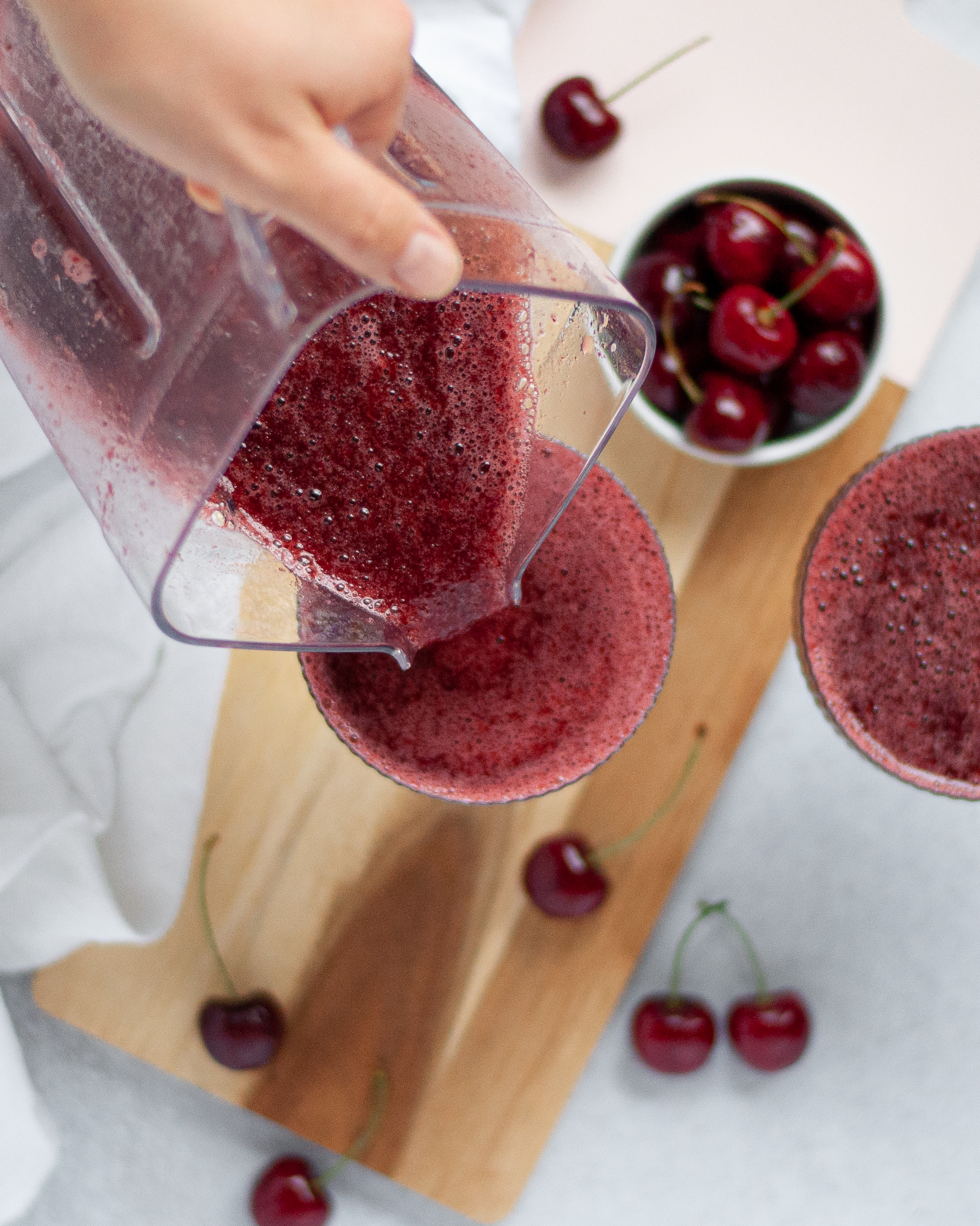 Top down view of a cocktail glass being filled with cherry frose from the blender pitcher. The glass is sitting on a wooden board and surrounded by fresh cherries.