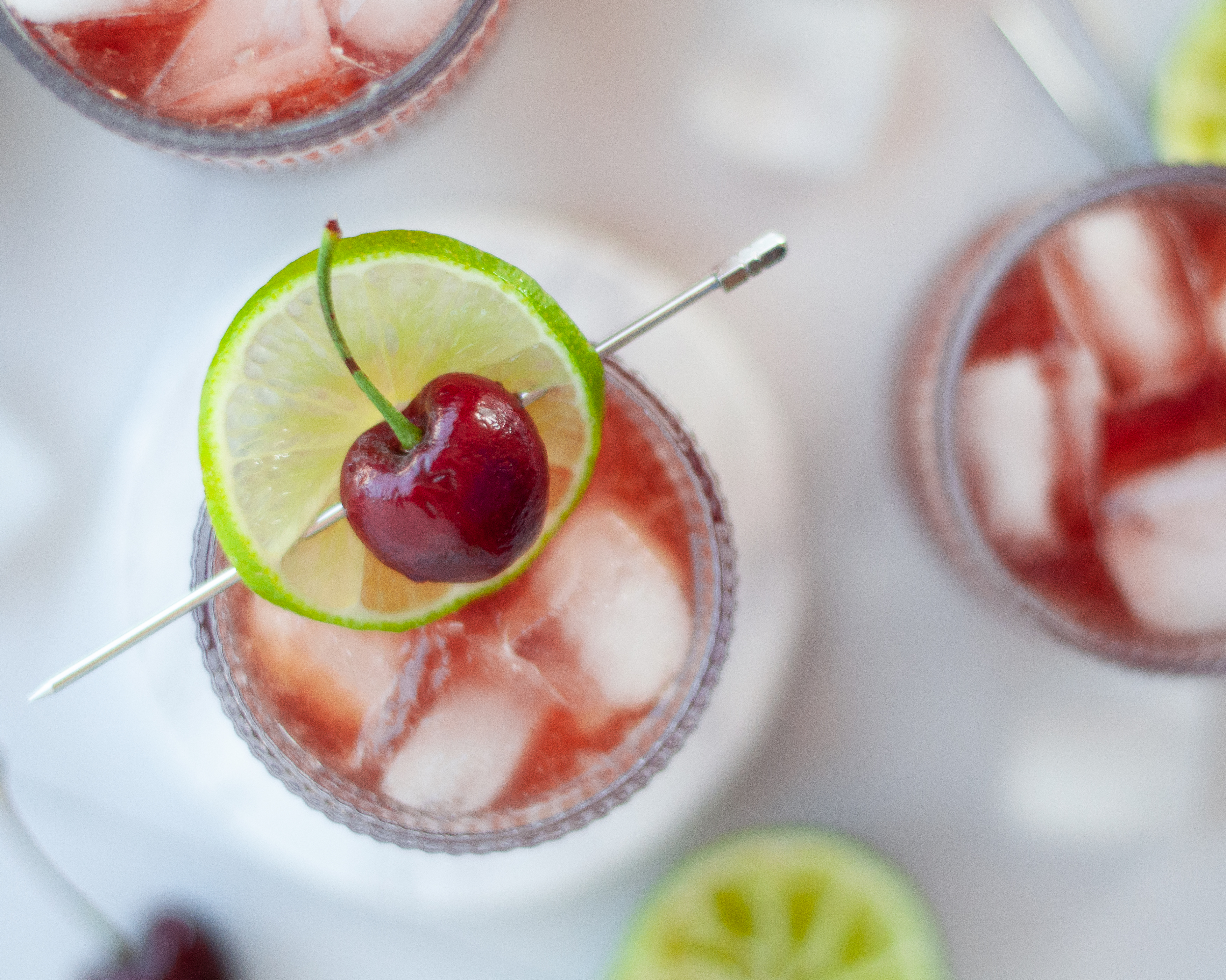Top down view of a glass of this cherry mocktail. The garnish of a fresh cherry and lime slice really jump out at you in this image. Surrounding the main glass is ice, pieces of lime, fresh cherries, and additional glasses filled with this easy non alcoholic drink recipe.