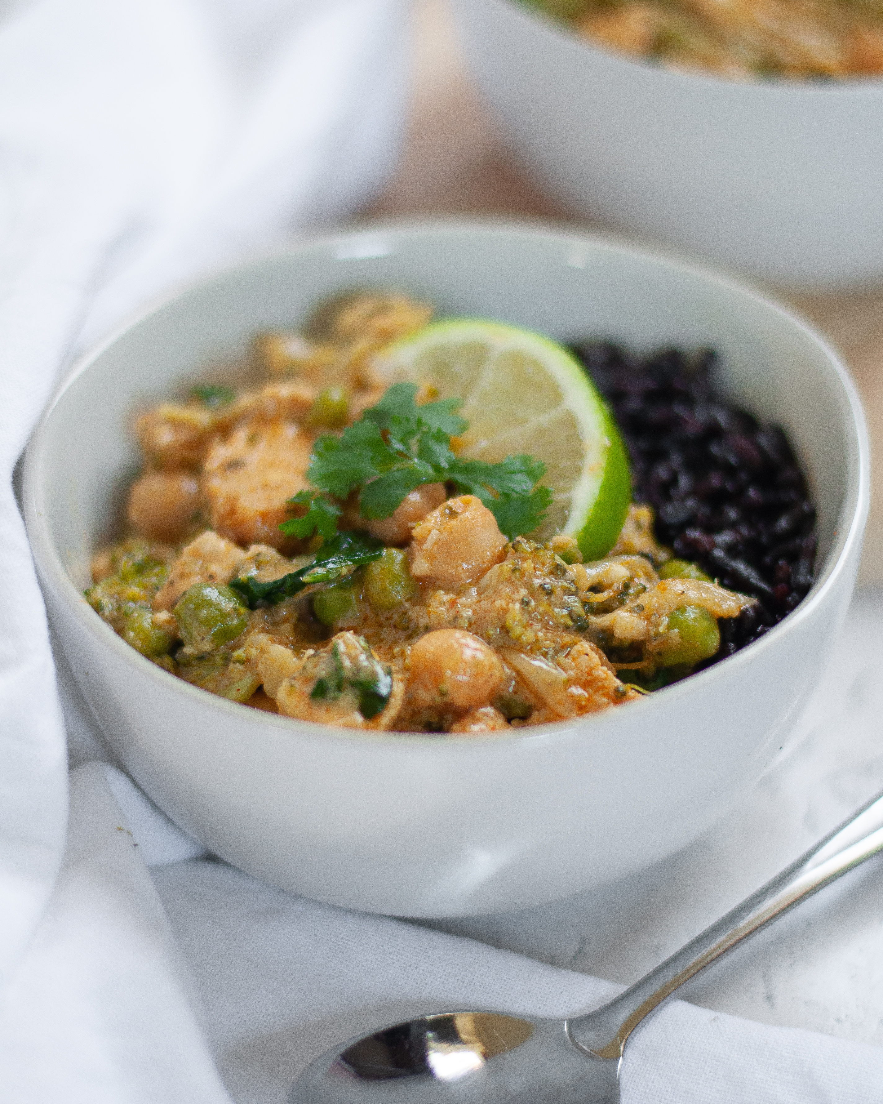 Close up shot of a bowl of chicken vegetable curry with chickpeas served with forbidden rice, a lime wedge, and cilantro.