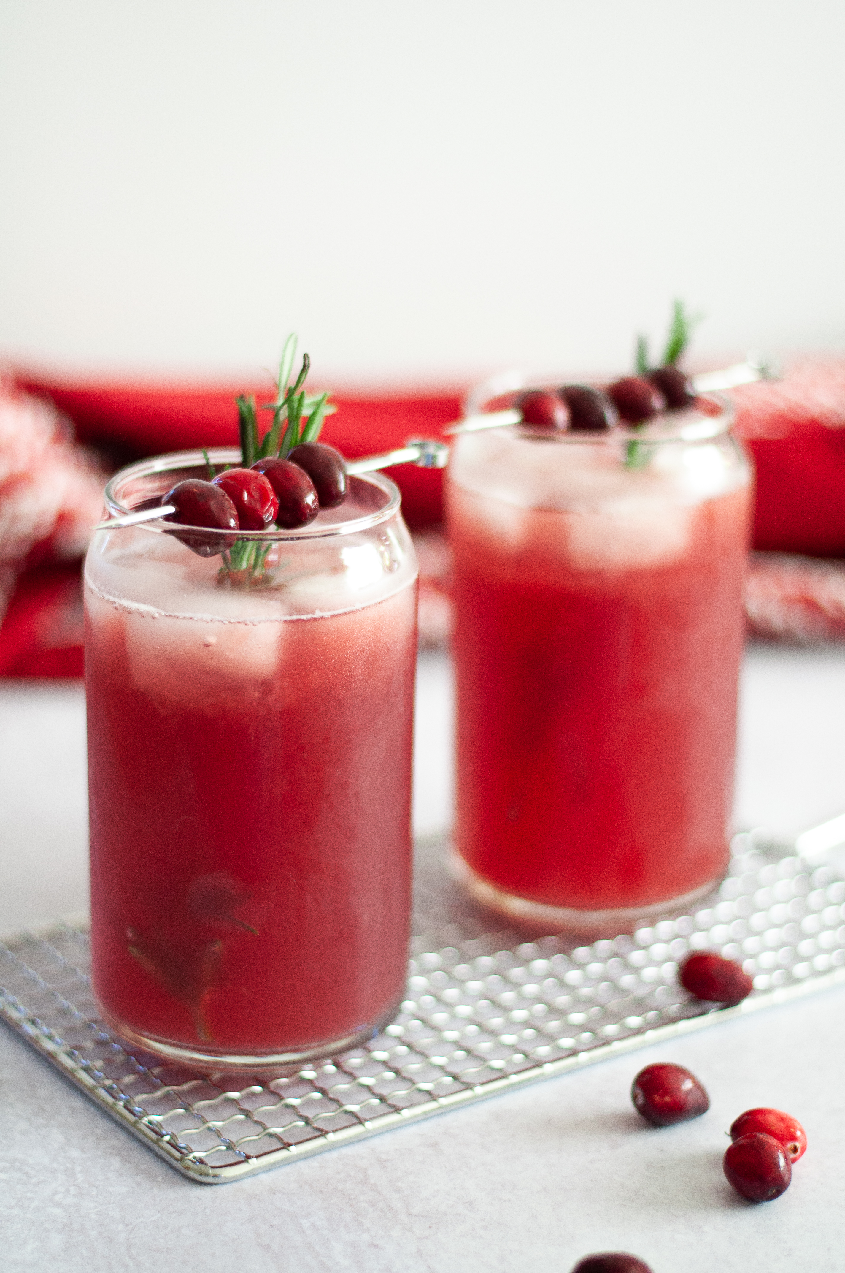 A different view of two glasses filled with this cranberry ginger mocktail, served on ice with a sprig of rosemary and a cocktail pick of cranberries