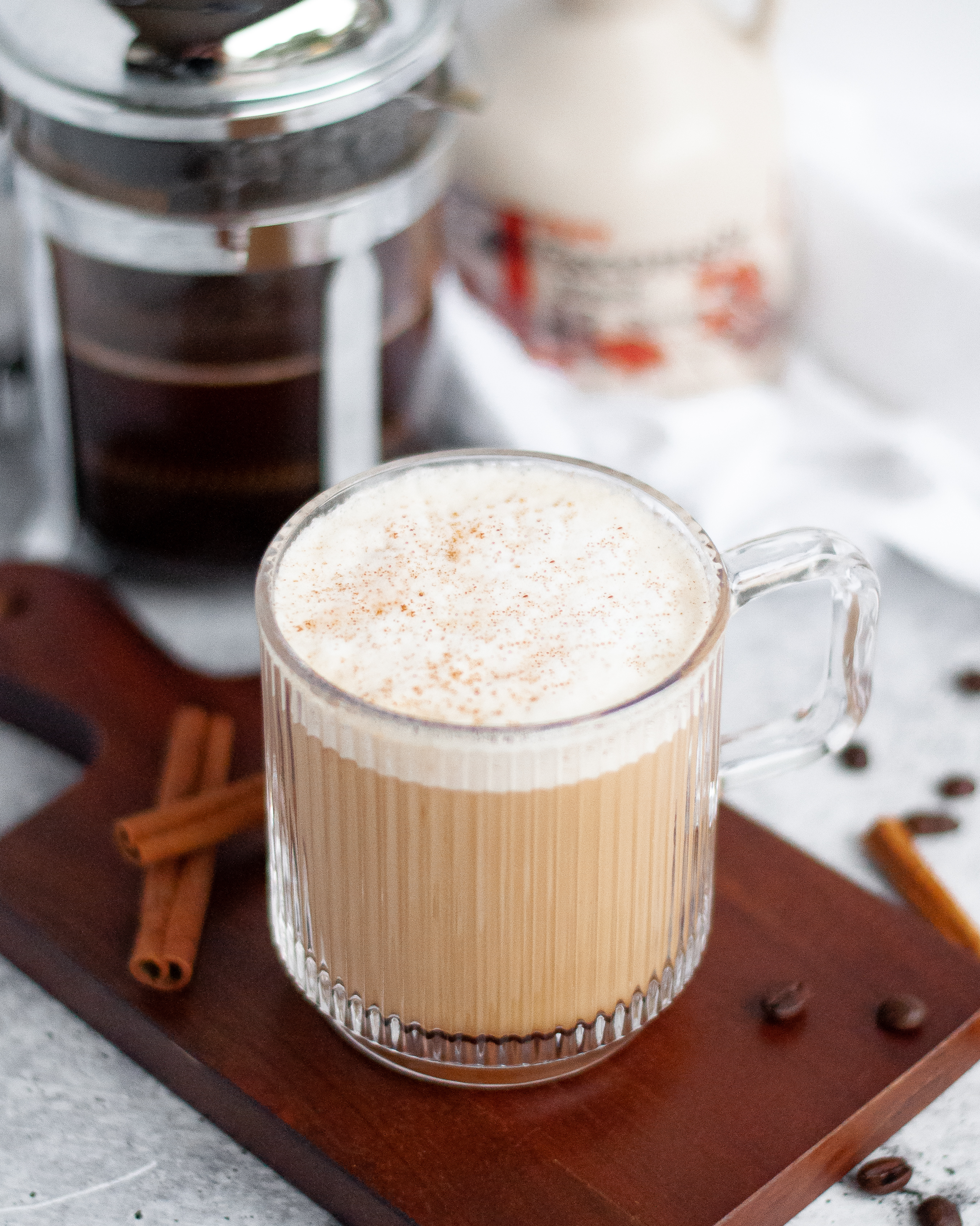 Glass mug filled with a hot maple cinnamon coffee with collagen. The maple cinnamon latte is sitting on a wooden board with coffee beans, cinnamon sticks, a jug of maple syrup, a french press, and a white napkin surrounding it.