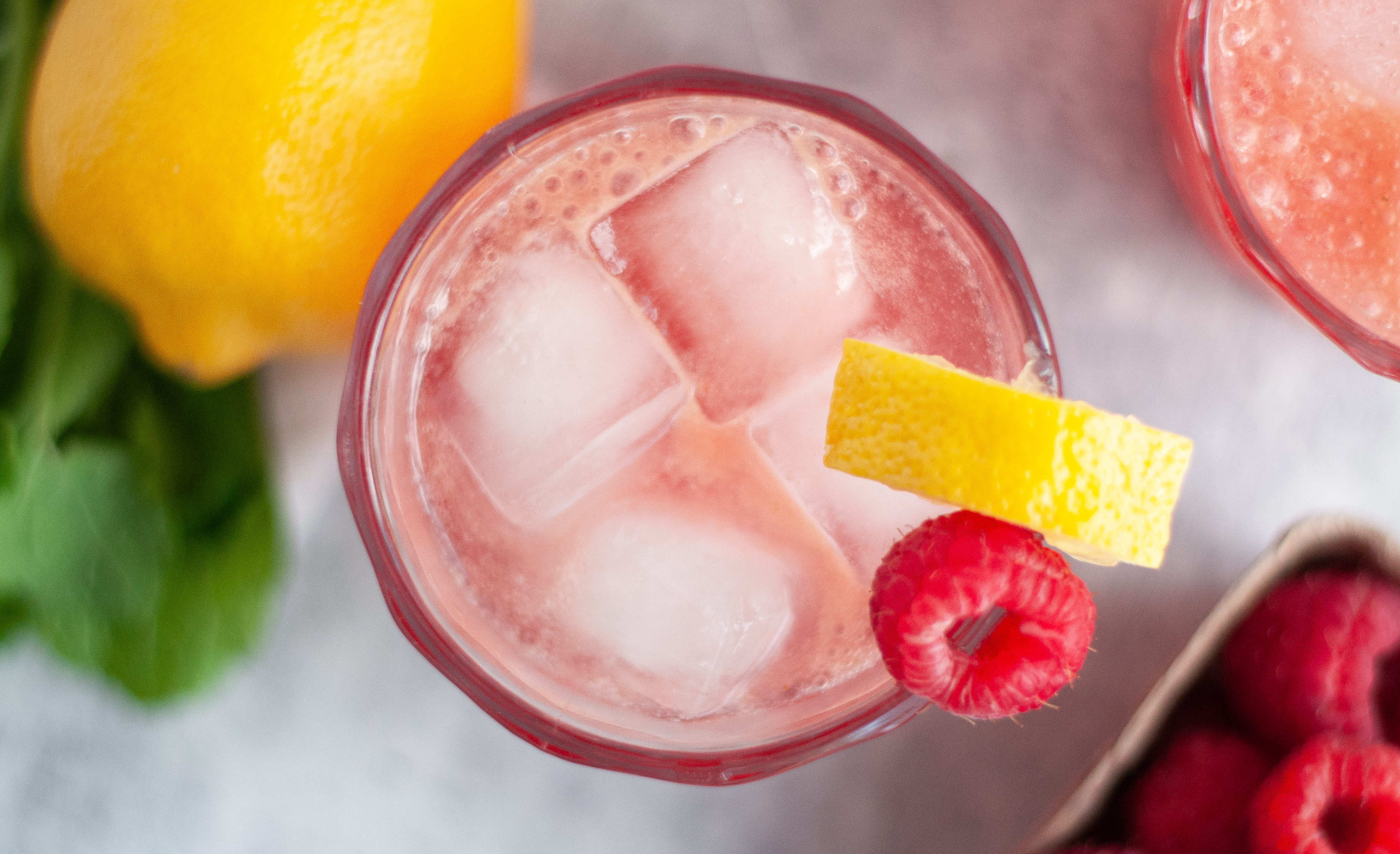 Top down view of a raspberry bourbon smash cocktail, garnished with a lemon wedge and raspberry. Surrounded by additional ingredients.