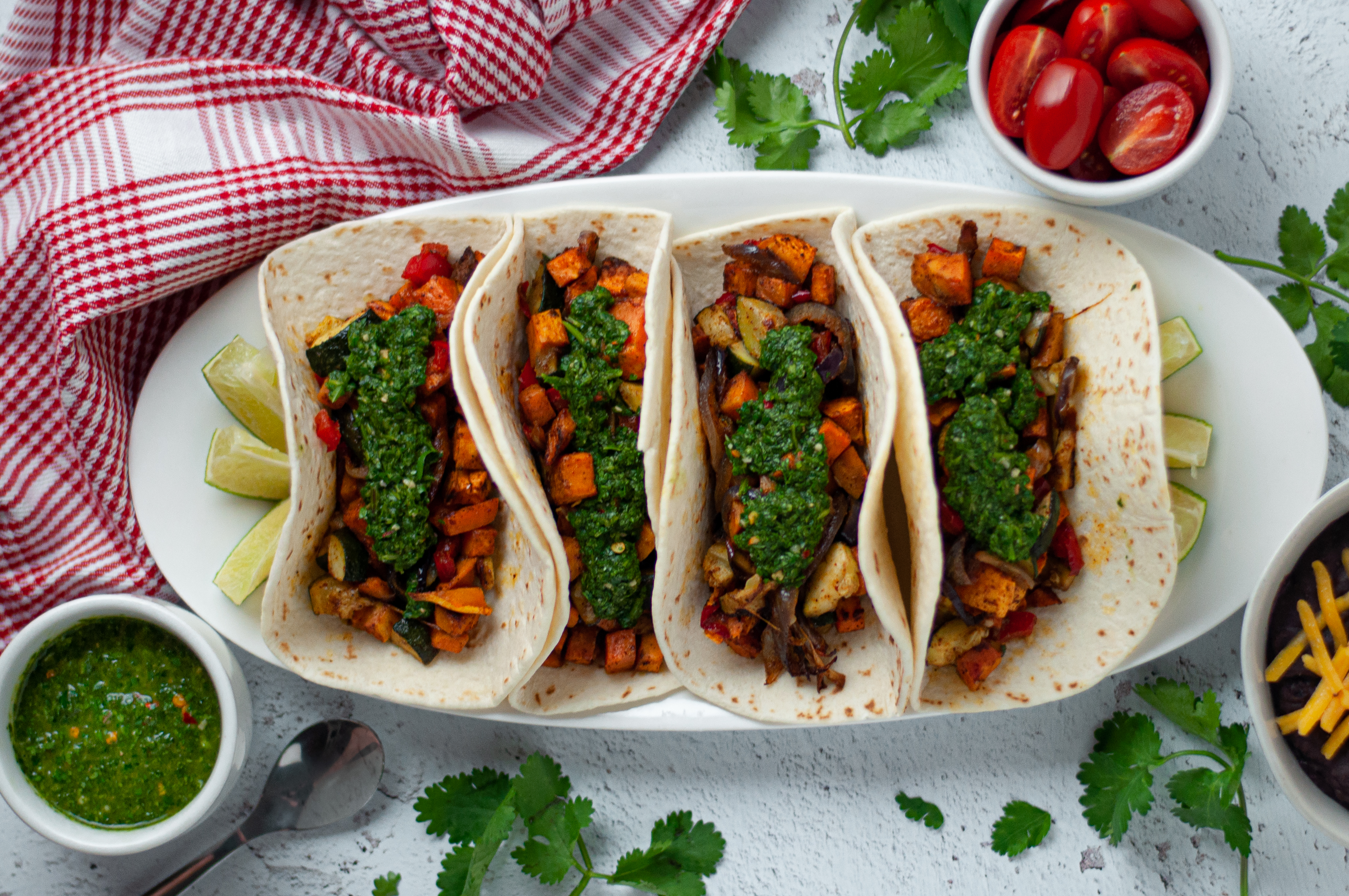 Top down view of a serving platter with 4 vegetarian tacos served up and ready to eat. Surrounded by a brightly colored napkin, tomatoes, cilantro, chimichurri sauce, and a side of beans