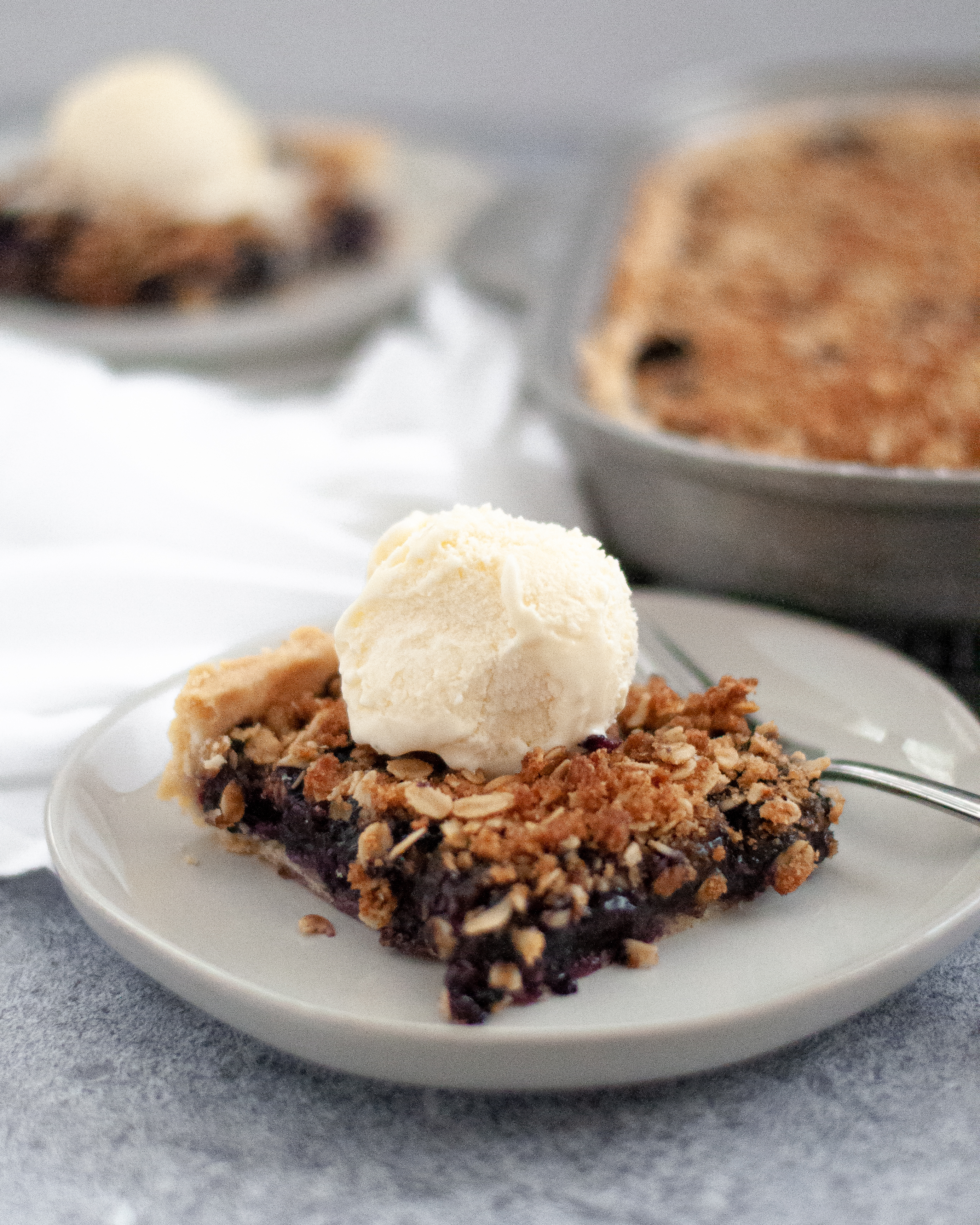 Close up of a slice of these blueberry pie bars. In the background you can see the pan holding the rest of the bluebery slab pie and another slice of blueberry pie bars on a plate. Each slice of this blueberry slab pie has a scoop of ice cream on top.