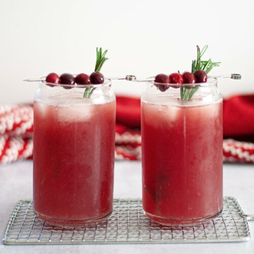 close up of two tall glasses filled with this cranberry ginger mocktail. the glasses are garnished with sprigs of fresh rosemary and cocktail picks with fresh cranberries.