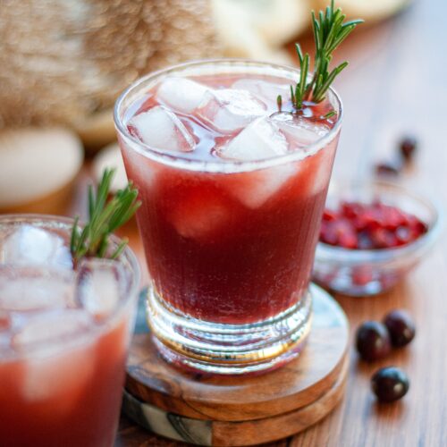 two glasses filled with this pomegranate and cranberry mocktail recipe. the glasses are garnished with a sprig of fresh rosemary and surrounded by fresh cranberries, pomegranate seeds, and gold decor.