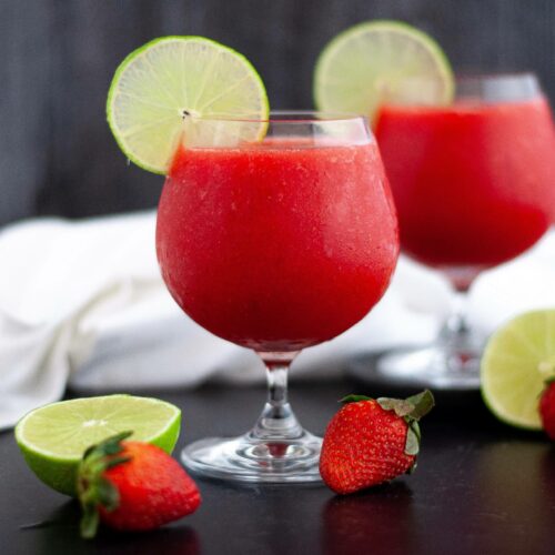 close up of a glass of bright red frozen strawberry daiquiri garnished with a lime slice.