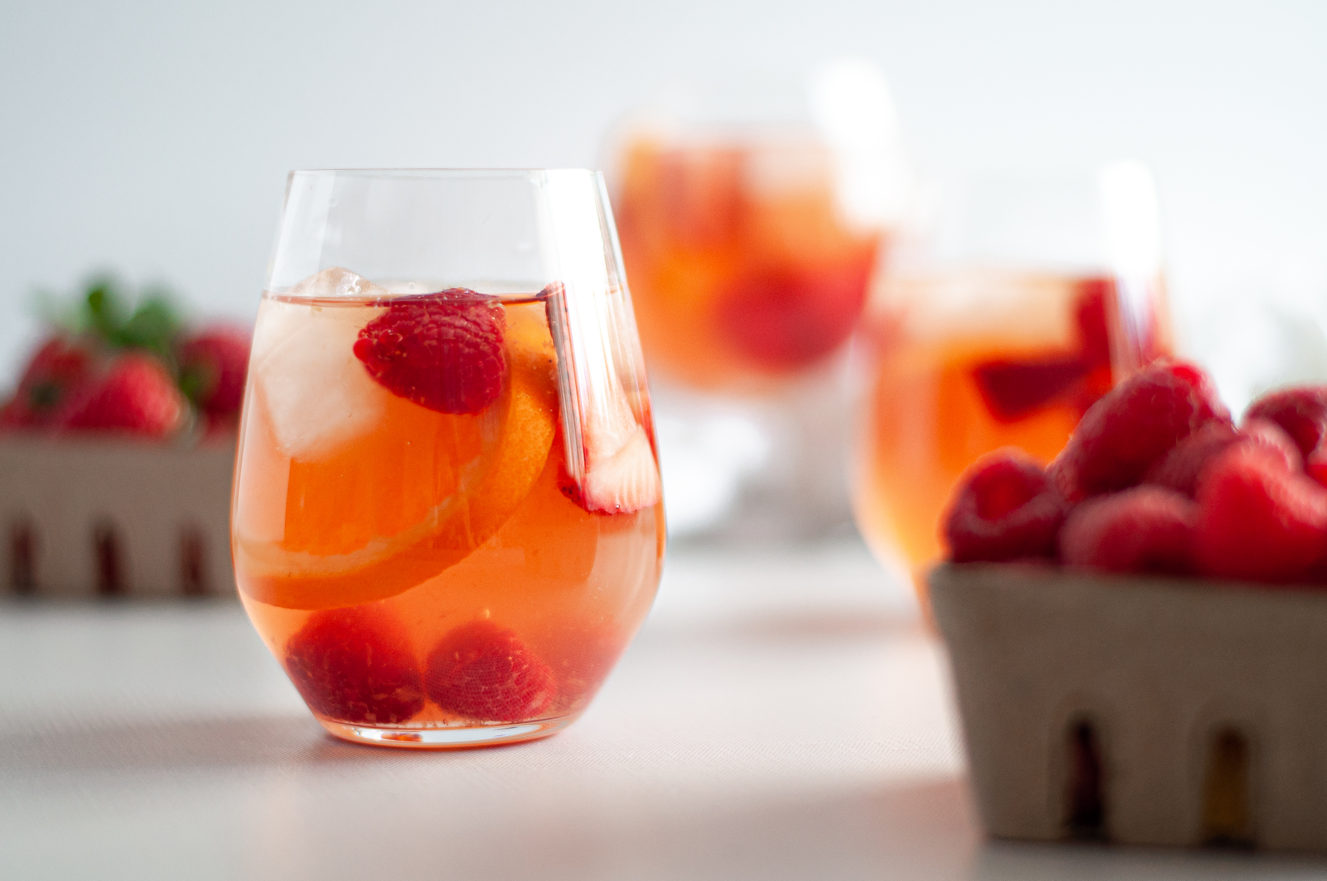 Straight on view of a stemless wine glass filled with rosé sangria, berries, orange slices, and ice. Surrounded by containers of berries and additional glasses filled with this rosé sangria recipe.