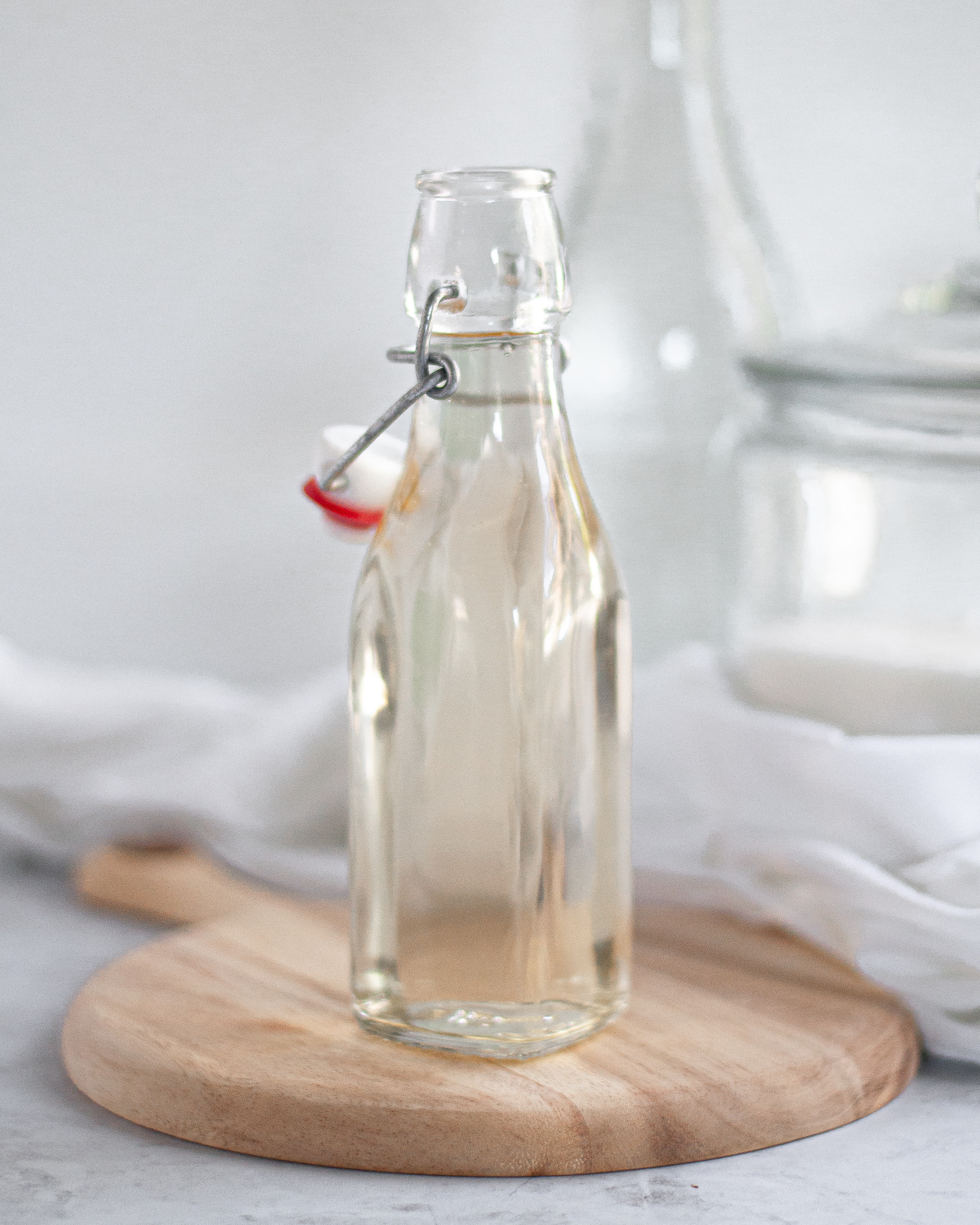 Container of simple syrup on a wooden block, with a napkin, jug of water, and container of sugar in the background.