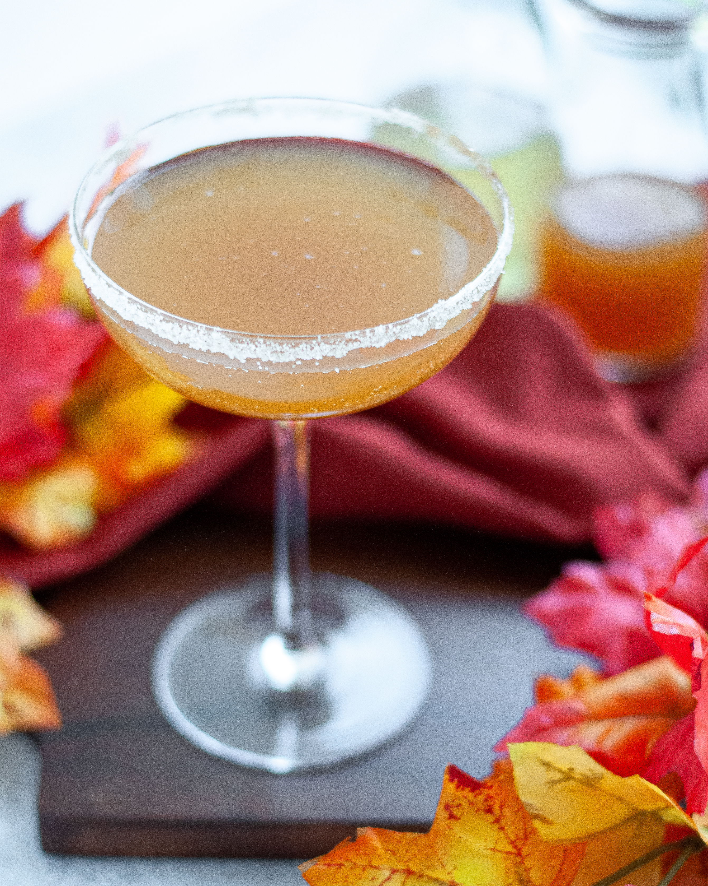 close up of a coupe glass filled with an apple cider mocktail with a sugar rim. The glass is surrounded by fall leaves, an orange linen, and glass jars with ginger beer and apple cider.