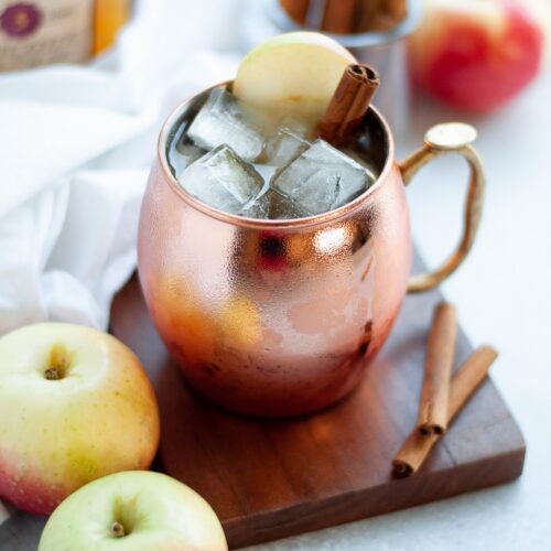 copper mug filled with this Apple Cider Bourbon Mule recipe and garnished with a cinnamon stick and apple slice. additional apples and cinnamon sticks surround the mug.