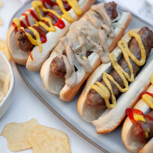 close up of a serving tray covered with beer brats on buns and topped with a variety of toppings like donions, mustard, and ketchup.