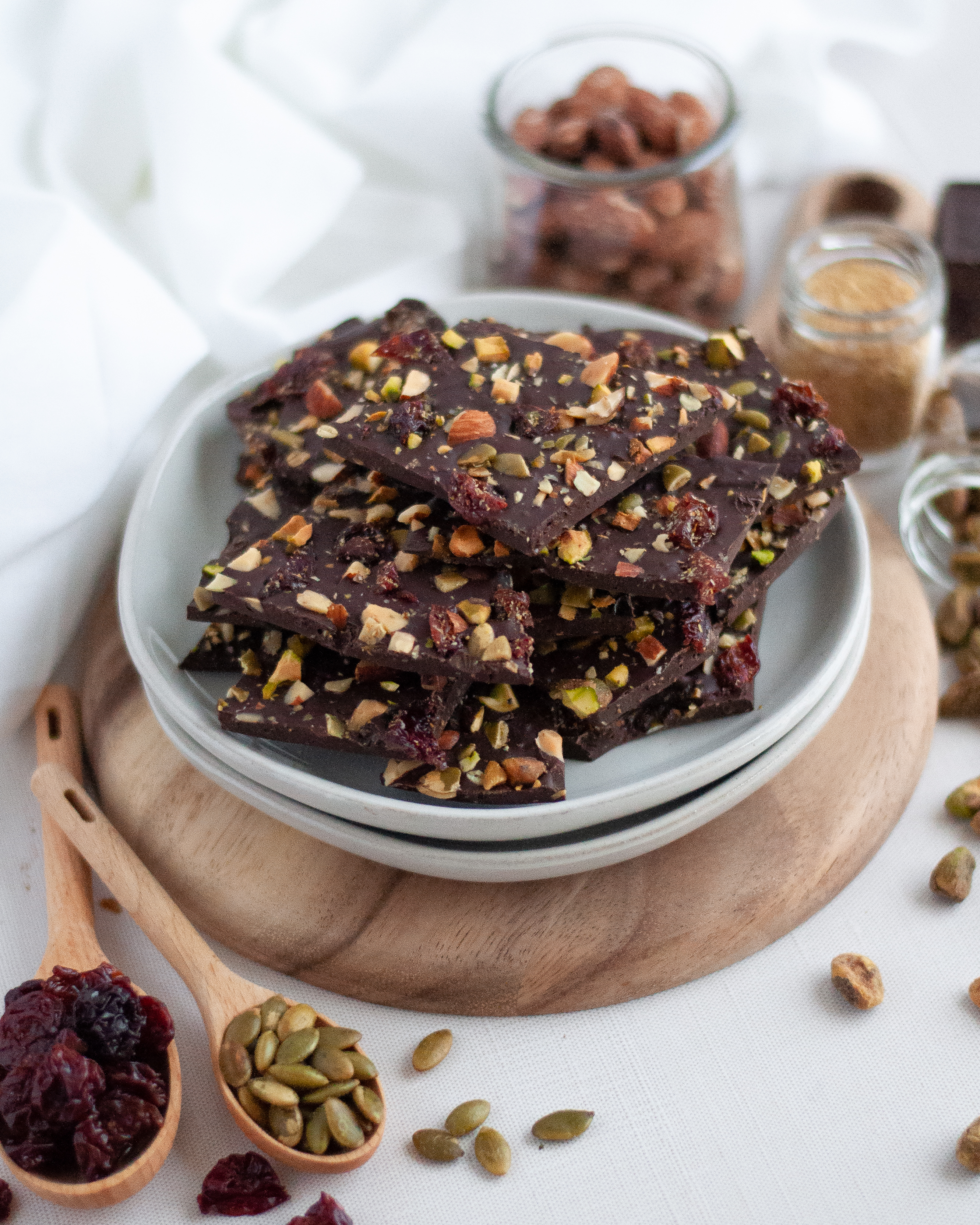 Side view of a plate stacked with dark chocolate bark with fruit and nuts, surrounded by the ingredients for chocolate bark: chocolate, ground flax, almonds, pistachios, pepitas, and cherries.
