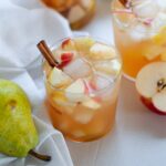low ball glass filled to the brim with white wine sangria with vodka. The glass is garnished with a cinnamon stick and surrounded by fresh fruit and more sangria glasses.