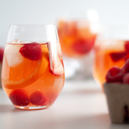 close up of a stemless wine glass filled with rose berry sangria. additional glasses are in the background, and cardboard containers of fresh berries surround the glass.