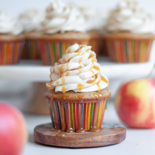 close up of an apple spice cupcake topped with spiced buttercream frosting, drizzled with caramel. The main cupcake is surrounded by fresh apples and additional cupcakes.