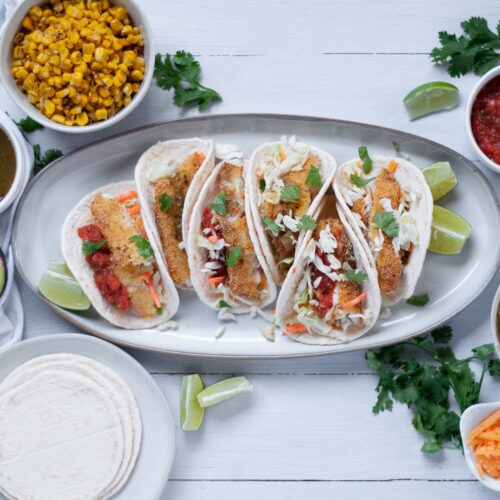 a serving tray filled with Cornmeal Crusted Fish Tacos and surrounded by bowls filled with a variety of sides and toppings.