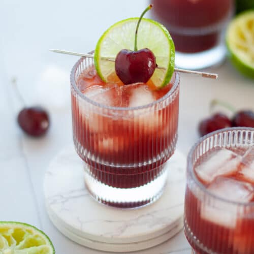 The focus of this image is on the center glass, filled with this lime and cherry mocktail and ice, and garnished with a slice of lime and cherry on a cocktail pick. The glass sits on a two white coasters, and there are additional glasses filled with this easy non alcoholic drink in the foreground and background. The glass is also surrounded with juiced limes, cherries, and ice.