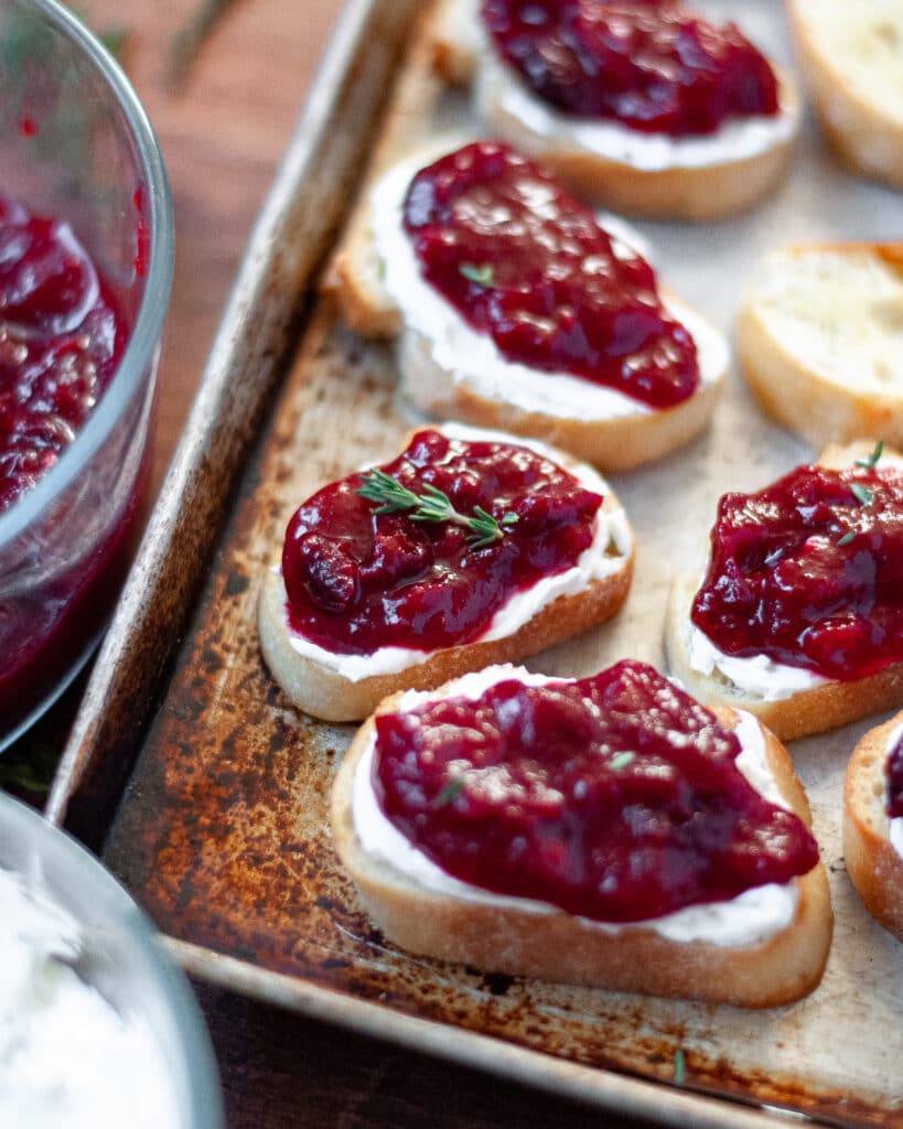 Baking sheet full of prepared cranberry and goats cheese crostini. There are bowls of whipped goat cheese with thyme and fresh cranberry sauce peaking into the side of the image.