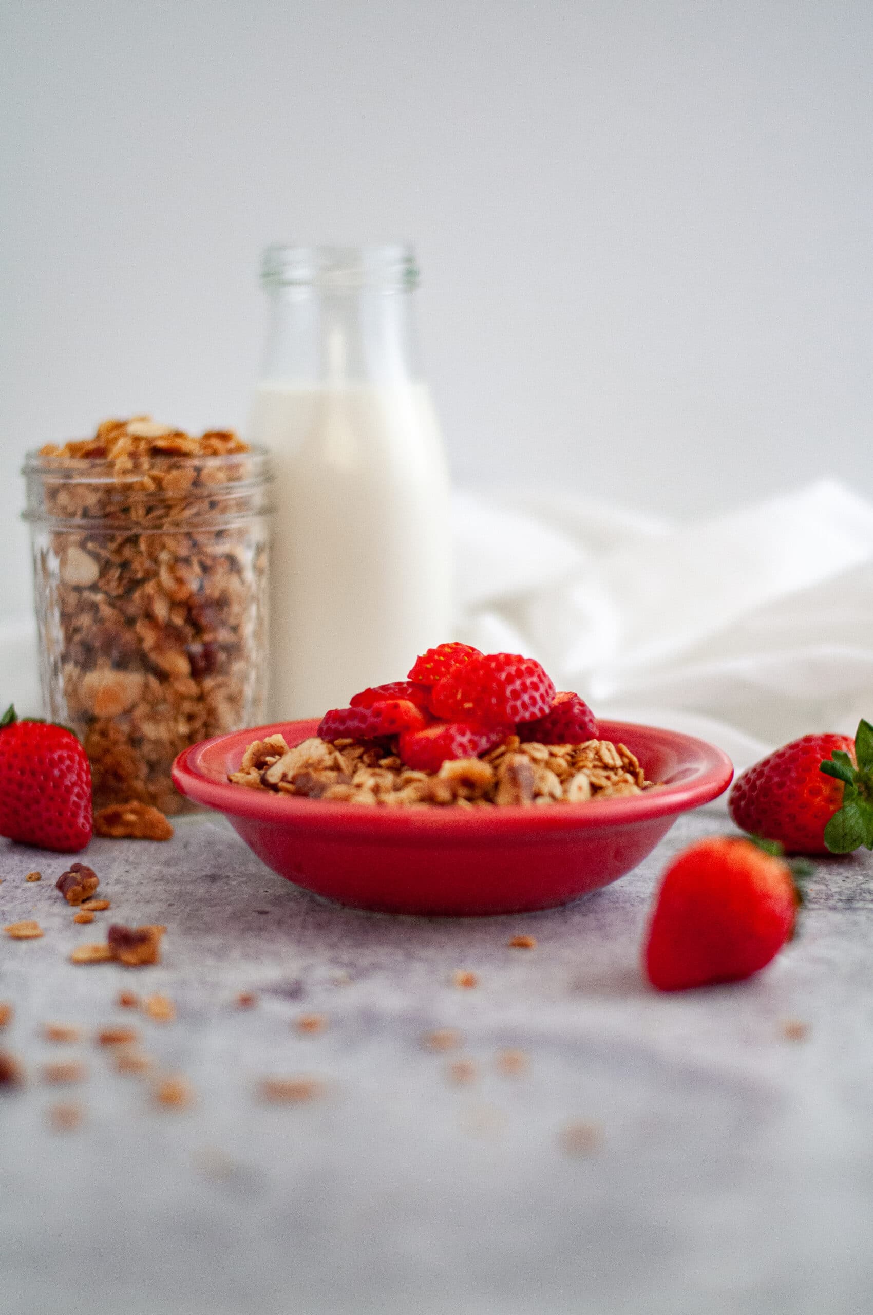 Small red bowl of granola topped with slices of strawberries. This homemade granola is surrounded by strawberries, granola in a small glass jar as well as scattered around, and a glass jar of milk