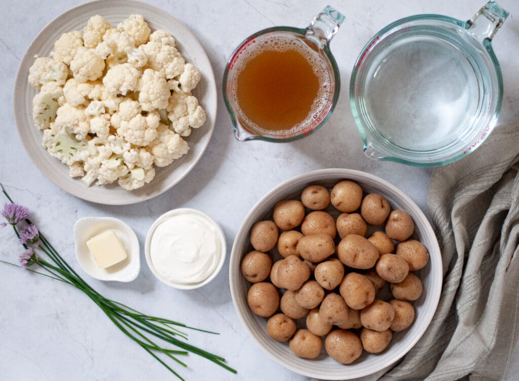 Ingredient show for these healthy mashed potatoes. Includes plates/dishes of raw cauliflower, baby gold potatoes, broth, water, butter, sour cream, and fresh chives.