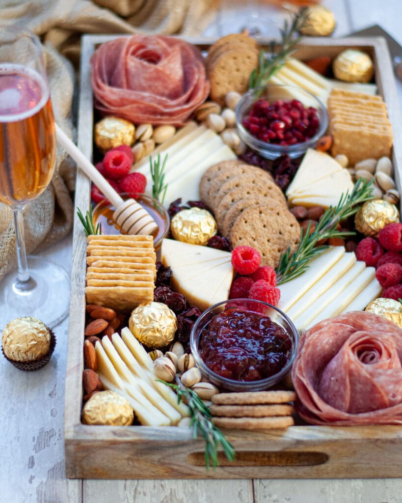 View of a wooden tray filled and beautifully displayed with all the ingredients for this new years eve charcuterie board. There is a glass of champagne next to the board and a golden linen in the background.